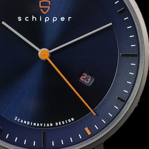 Puede incluir: Primer plano de una esfera de reloj azul con agujas blancas y detalles en naranja. La esfera del reloj tiene el texto "schipper" y "SCANDINAVIAN DESIGN". La ventana de fecha muestra el número "23".