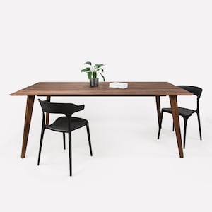 Könnte beinhalten: Ein Esstisch aus Holz mit brauner Oberfläche und vier schwarzen Beinen. Zwei schwarze Stühle stehen auf beiden Seiten des Tisches. Eine kleine Topfpflanze steht auf dem Tisch.