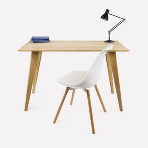 Op de afbeelding: Een witte stoel met houten poten staat voor een houten bureau met een zwarte bureaulamp. Een stapel blauwe boeken ligt op het bureau.
