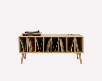 Pode incluir: Um suporte de toca-discos de madeira clara com 10 ranhuras para discos de vinil. O suporte tem uma plataforma giratória na parte superior e quatro pernas.