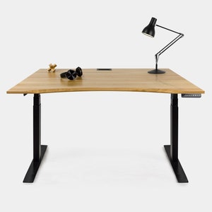 Pode incluir: Uma mesa de pé ajustável preta com tampo de madeira. A mesa tem uma borda curva e um painel de controle preto no lado direito. Há uma lâmpada, fones de ouvido e um pequeno objeto de madeira na mesa.
