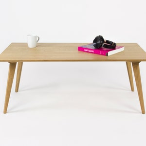 Może przedstawiać: Jasny drewniany stolik kawowy z czterema nogami. Biały kubek i książka w różowej okładce oraz czarne słuchawki leżą na blacie.