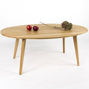 Peut inclure: Une table basse ovale en bois clair avec quatre pieds effilés. La table a une finition en bois naturel et est décorée de deux pommes rouges et d'une branche de fleurs séchées.