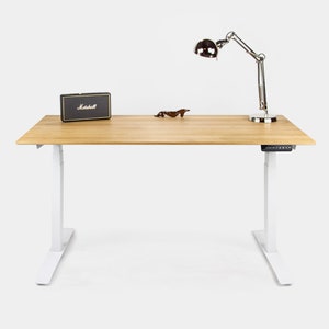 Peut inclure: Un bureau debout blanc avec un plateau en bois. Le bureau a un panneau de commande noir sur le côté droit et une lampe de bureau argentée sur le côté gauche. Il y a une petite boîte noire avec le mot "Marshall" dessus sur le bureau.