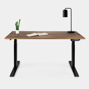 Peut inclure: Un bureau debout réglable noir avec un plateau en bois de noyer. Le bureau a une petite plante en pot et une lampe noire sur la surface.