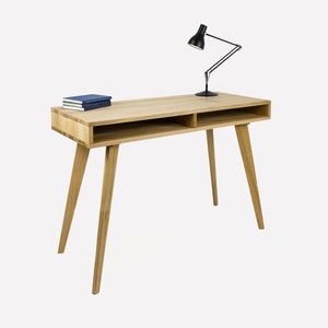 Puede incluir: Un escritorio de madera clara con dos estantes y patas cónicas. Una lámpara de escritorio negra está sobre el escritorio con una pila de libros azules.