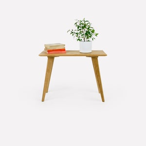 Op de afbeelding: Een houten bijzettafel met een lichtbruine afwerking en vier poten. Een kleine plant in een pot met groene bladeren en een witte pot staat op de tafel, samen met een stapel boeken.