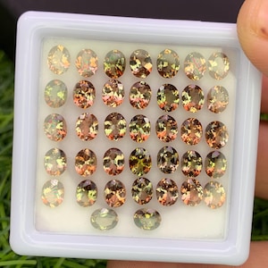May include: A collection of oval-cut gemstones in a white plastic tray. The gems display a mix of colors, including shades of brown, green, and orange. The gemstones are arranged neatly, showcasing their polished surfaces and internal patterns.