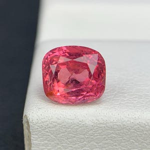 May include: A cushion-cut gemstone with a vibrant, saturated pinkish-red hue. The stone features a faceted surface that reflects light, creating a brilliant appearance. It is positioned on a textured white surface with a black background visible.