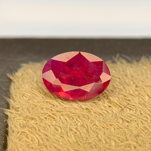 Könnte beinhalten: Ein leuchtender, oval geschliffener Edelstein in tiefroter Farbe mit einer facettierten Oberfläche, die das Licht reflektiert. Der Stein weist eine symmetrische Anordnung von dreieckigen und trapezförmigen Facetten auf der Oberseite auf.