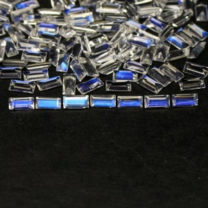 Könnte beinhalten: Eine Sammlung von klaren, facettierten, rechteckigen Edelsteinen mit einem blauen, irisierenden Schimmer. Die Edelsteine sind auf einer schwarzen Oberfläche verstreut.