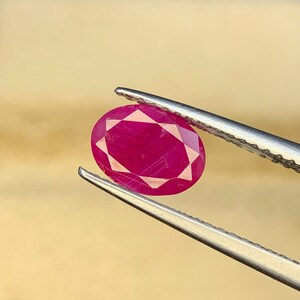 May include: A vibrant, oval-cut ruby gemstone with a deep pinkish-red hue and faceted surface, held securely by a pair of silver-toned metal tweezers. The gemstone displays internal clarity and light reflection across its polished, symmetrical facets.