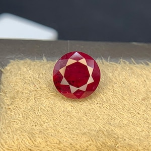 May include: A round, faceted, deep red gemstone featuring a prominent eight-pointed star pattern cut into its surface. The stone displays a vibrant crimson hue with sharp, geometric light reflections across its polished, symmetrical facets.