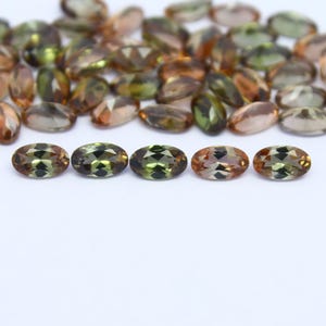 May include: An assortment of oval-cut gemstones in shades of green, brown, and amber. The gemstones are scattered across a white surface, with a few in sharp focus at the bottom. These faceted stones are ideal for jewelry making.