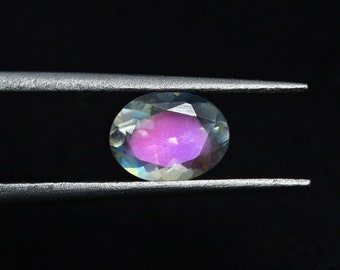 May include: A close-up of a iridescent oval gemstone with a pink hue, held in a pair of tweezers against a black background.