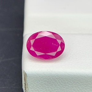 Pode incluir: Uma gema de rubi facetada em forma oval. A gema é de uma cor vermelho-rosada vibrante e profunda, com múltiplas facetas que capturam a luz. O rubi está colocado sobre um fundo branco.