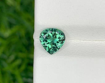 May include: A heart-shaped green gemstone with a faceted cut. The gemstone is set against a white background.
