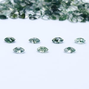 Puede incluir: Siete piedras preciosas de ágata verde musgo en forma de marquesa, con un aspecto claro y translúcido. Las piedras están dispuestas en fila sobre una superficie blanca.