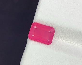 Puede incluir: Una gema rectangular de color rosa intenso con bordes biselados. La gema se encuentra sobre una superficie blanca texturizada, con un fondo oscuro. El color de la gema es un rosa audaz y saturado.