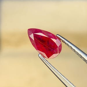 Puede incluir: Una piedra preciosa roja tallada en forma de pera con facetas brillantes, sostenida por unas pinzas de metal plateado. La piedra muestra tonos carmesí profundos y claridad interna con facetas visibles que reflejan la luz.