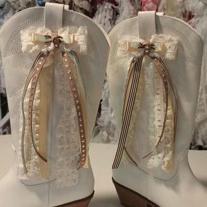May include: Pair of white cowboy boots with decorative lace and ribbon accents. The boots feature a light brown heel and are adorned with cream-colored lace, ribbons, and star-studded embellishments. The boots are displayed against a blurred background.