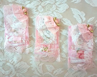 May include: Three pink and white lace sachets with floral accents. Each sachet features layers of delicate lace, a pink fabric base, and a decorative floral embellishment. The sachets are arranged on a white lace tablecloth.