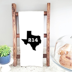 May include: White kitchen towel with a black silhouette of the state of Texas and the number 214 printed inside the state. The towel is hanging on a wooden ladder.