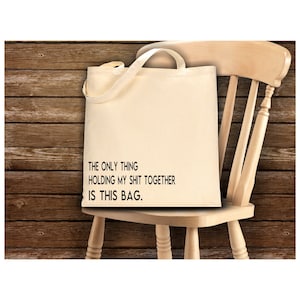 May include: A beige canvas tote bag with the text "THE ONLY THING HOLDING MY SHIT TOGETHER IS THIS BAG." printed on it. The bag is sitting on a wooden chair.