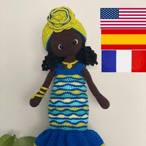 Puede incluir: Muñeca de ganchillo hecha a mano con piel oscura, vestida con un vestido azul y amarillo. La muñeca tiene cabello negro rizado, un turbante amarillo y una pulsera a juego. El vestido presenta una falda con volantes y un patrón ondulado en el corpiño. La muñeca mide unos 30 cm de alto.