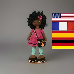 May include: A handcrafted crochet doll with dark skin, black curly hair, and a pink dress. The doll has a small black handbag with a yellow tassel and is wearing blue leggings and brown shoes. A pink flower adorns the doll's hair.