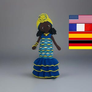 May include: Crocheted doll with dark skin, wearing a blue and yellow dress with a ruffled skirt. The doll has a yellow head wrap with a flower and black curly hair. A small gold necklace and bracelet complete the look.