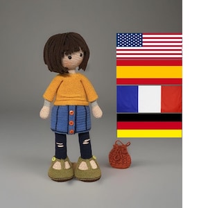 Puede incluir: Una muñeca de ganchillo con pelo de lana marrón, que lleva una camiseta amarilla de manga larga, una falda azul con tres botones naranjas y unos leggings azul oscuro con detalles de puntadas horizontales claras. Lleva zapatos verde oliva.