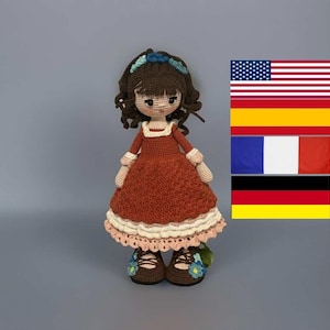 Könnte beinhalten: Handgefertigte Häkelpuppe mit braunen Haaren, die ein rostfarbenes Kleid mit weißem Besatz und ein Blumenstirnband trägt. Die Puppe hat braune Schuhe mit blauen Blumenakzenten. Der Hintergrund ist grau.