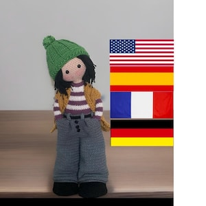 Könnte beinhalten: Handgemachte Häkelpuppe mit schwarzen Haaren, die eine grüne Mütze, ein gestreiftes Hemd, eine gelbe Weste und eine graue Hose trägt. Die Puppe hat schwarze Schuhe und einen neutralen Hautton. Flaggen der Vereinigten Staaten, Spaniens, Frankreichs und Deutschlands im Hintergrund.