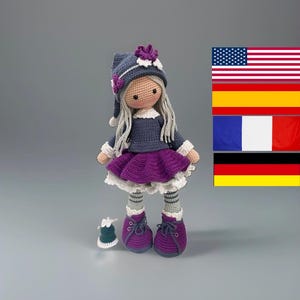 Könnte beinhalten: Eine handgefertigte Häkelpuppe mit langem grauem Garnhaar, die eine dunkelblaue Mütze mit einer violetten Blume, ein dunkelblaues Langarmshirt, einen gerüschten violetten Rock mit weißem Saum, grau gestreifte Leggings und violette Schnürschuhe trägt, begleitet von einer kleinen Glocke.