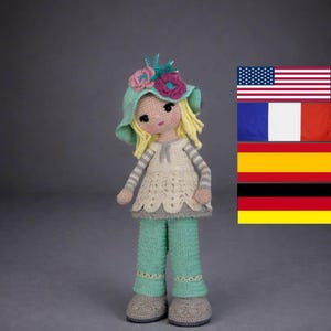 Könnte beinhalten: Eine handgefertigte Häkelpuppe mit einem hellgrünen Hut, der mit rosa und blauen Blumen verziert ist. Die Puppe hat blondes Haar, ein cremefarbenes Kleid und hellgrüne Hosen. Die Schuhe der Puppe sind grau. Die Puppe steht vor grauem Hintergrund.