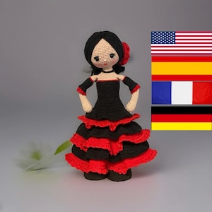 Op de afbeelding: Handgemaakte gehaakte pop in een flamenco danseres outfit. De pop heeft zwart haar, een rode bloem en draagt een zwarte jurk met rode ruches. Vlaggen van de Verenigde Staten, Spanje, Frankrijk en Duitsland op de achtergrond.