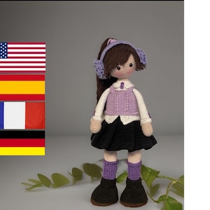 Könnte beinhalten: Eine handgefertigte Häkelpuppe mit braunen Haaren, die eine lila Weste, einen schwarzen Rock und schwarze Stiefel trägt. Die Puppe hat ein weißes Hemd unter der Weste und lila Ohrenschützer. Die Puppe steht auf einer weißen Oberfläche mit grünen Blättern.