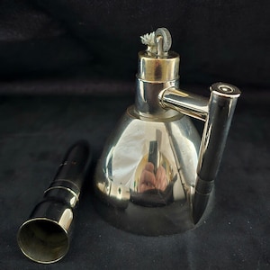 Könnte beinhalten: Eine versilberte Öllampe mit Docht und einem kleinen, runden, versilberten Behälter. Die Lampe hat einen Griff und einen Ausguss. Die Lampe steht auf einer schwarzen Oberfläche.