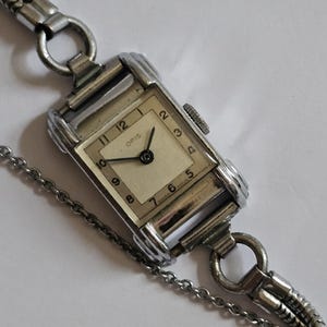 May include: A vintage Oris wristwatch with a rectangular silver-tone case and a cream face. The watch has black numerals and hands, and is attached to a silver chain bracelet. A classic timepiece, the watch is a vintage item.