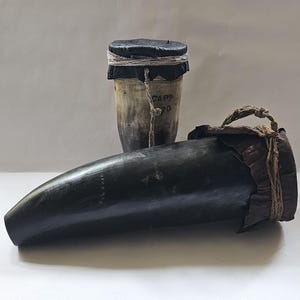 Könnte beinhalten: Zwei antike Musikinstrumente aus Tierhörnern. Eines ist ein dunkelbraunes Horn mit einer Leder- und Schnurummantelung. Das andere ist ein hellbraunes Horn mit einer schwarzen Lederkappe und einer Schnurwicklung. Das Horn mit der schwarzen Lederkappe trägt den Schriftzug "CAPP 70".