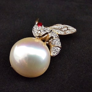 May include: A gold-tone bee brooch with clear rhinestones and a red centre stone, perched on a large, white pearl.