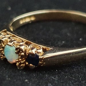 K&ouml;nnte beinhalten: Ein goldener Ring mit einem Opal und zwei blauen Saphiren, die in einem floralen Design gefasst sind.