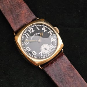 May include: Vintage Ingersoll watch with a gold-tone case and a black face. The watch features white numerals and hands, a subdial, and a brown leather strap. The watch has a rectangular shape with rounded corners.