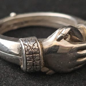 May include: A silver ring featuring two hands clasped together in a handshake. The ring has a detailed design, with the hands appearing to be holding each other. The band of the ring has an engraved pattern. The ring is displayed against a dark background.
