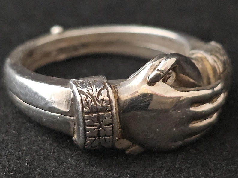 May include: A silver ring featuring two hands clasped together in a handshake. The ring has a detailed design, with the hands appearing to be holding each other. The band of the ring has an engraved pattern. The ring is displayed against a dark background.