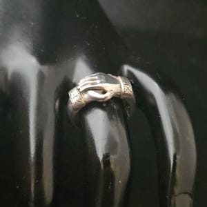 May include: A silver ring featuring a detailed design of two hands clasped together. The ring has a textured band with decorative elements. The hands are intricately sculpted, showcasing the ring's craftsmanship. The ring is displayed on a black surface.