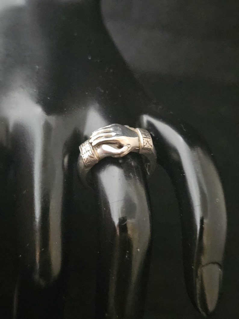 May include: A silver ring featuring a detailed design of two hands clasped together. The ring has a textured band with decorative elements. The hands are intricately sculpted, showcasing the ring's craftsmanship. The ring is displayed on a black surface.