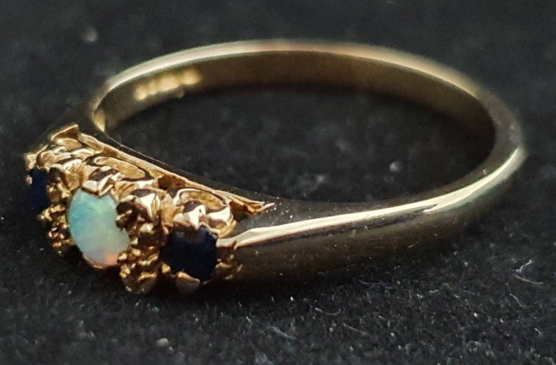K&ouml;nnte beinhalten: Ein goldener Ring mit einem Opal und zwei blauen Saphiren, die in einem floralen Design gefasst sind.