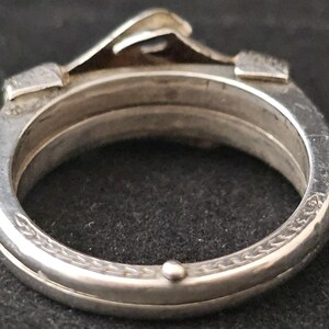 May include: Close-up of a silver ring with a unique design. The ring features a raised, architectural element and intricate detailing around the band. The ring is made of a shiny, metallic material, possibly sterling silver.